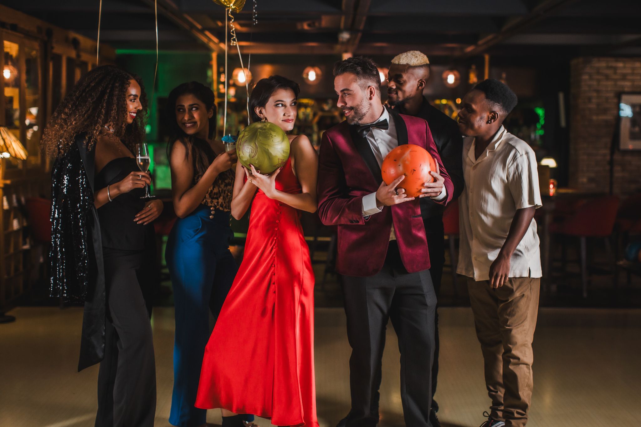 A diverse group of adults dressed elegantly in an bowling alley holding a bowling ball.