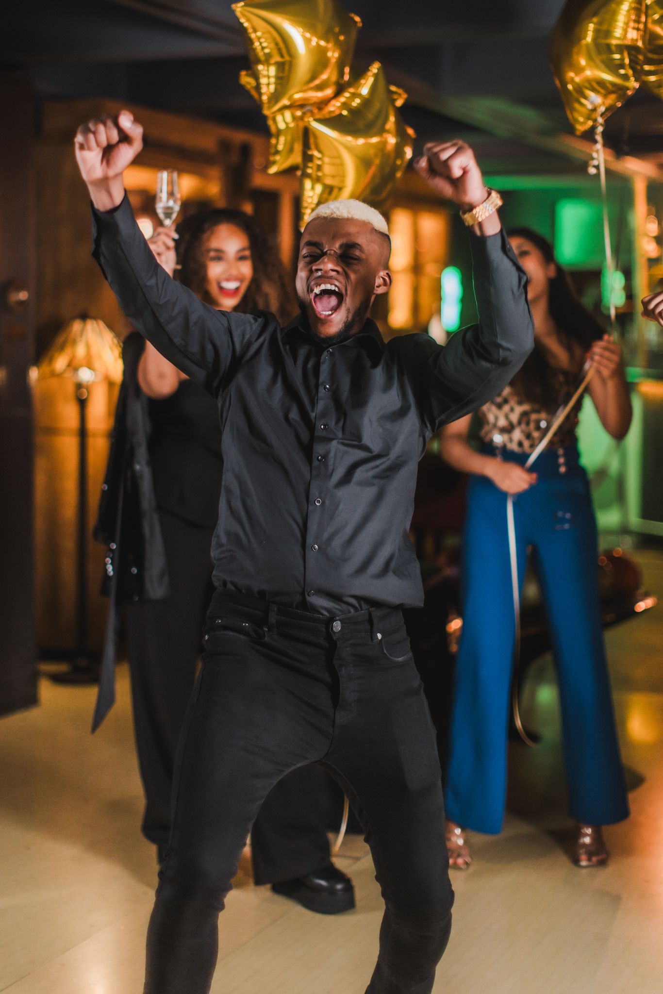 A man in a black shirt and pants joyfully dances among floating gold balloons.