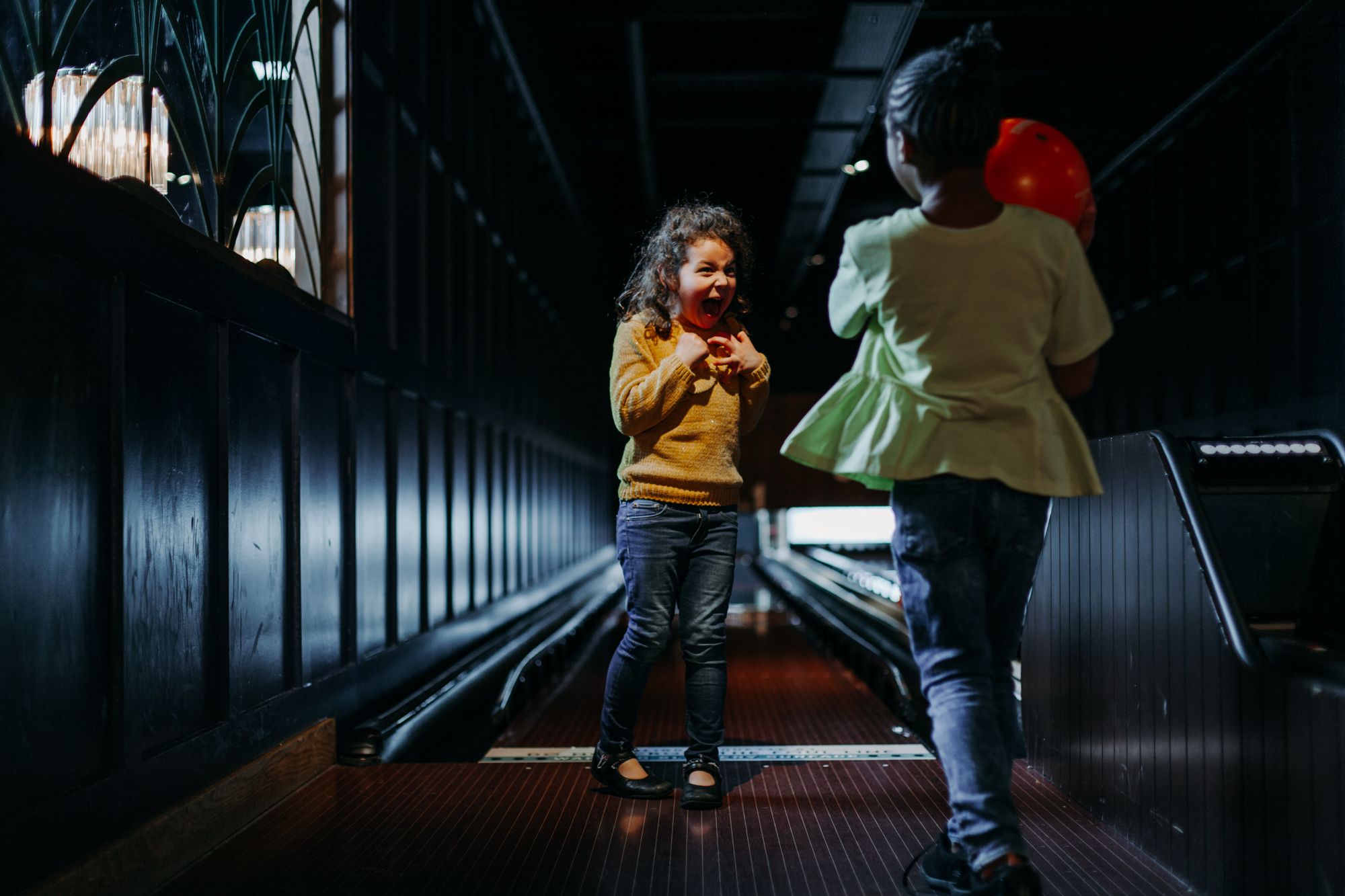 Two children stand in a dimly lit bowling alley, holding an orange bowling ball.