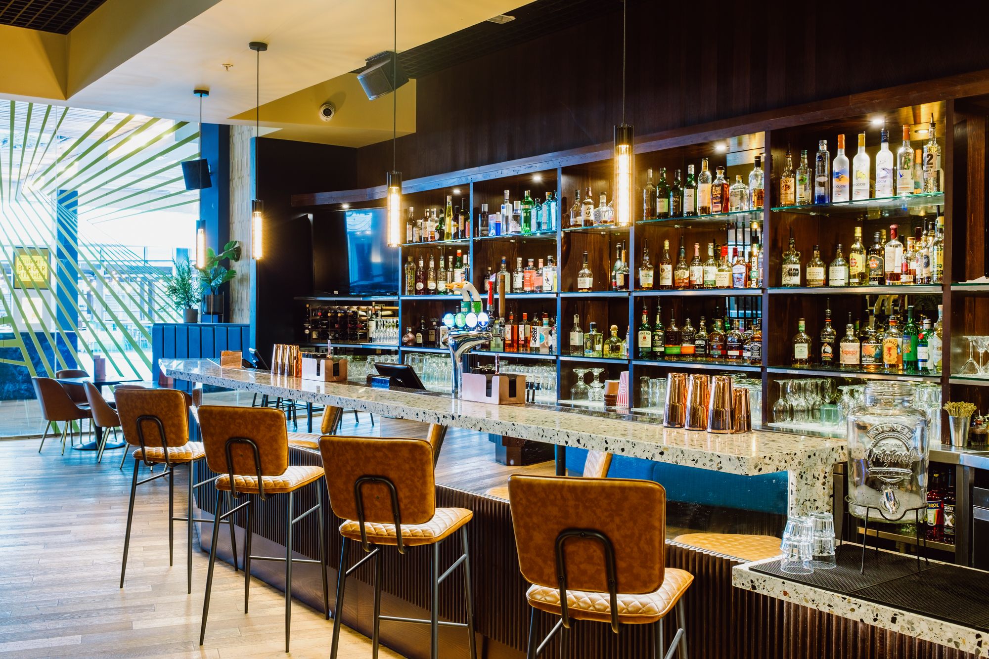 A bar showing many different bottles of alcohol with a table and bar stools.